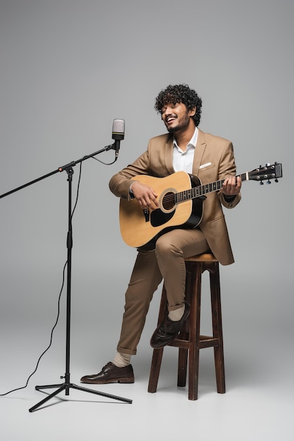 Un anfitrión indio sonriente con ropa formal tocando la guitarra acústica y cantando en el micrófono en el puesto