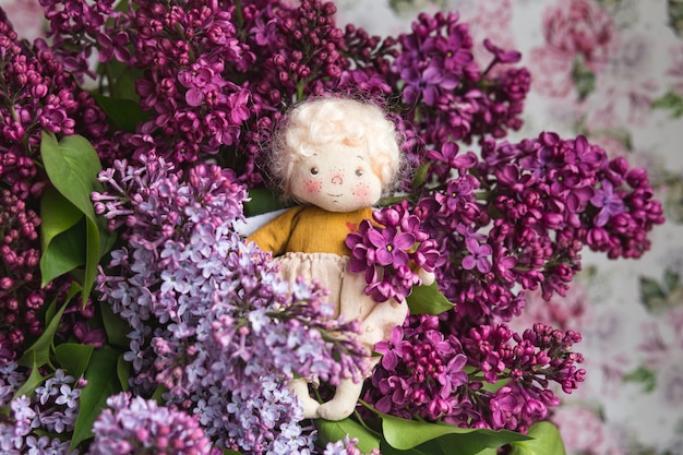 Angelito de cabello dorado en las flores de color azul, rosa, violeta, lila violeta. Juguete hecho a mano en colores violetas lilas. Tarjeta de felicitación.