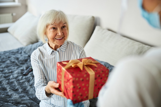 Angenehm überraschte alternde Person, die eine Schachtel in einem Geschenkpapier von einer häuslichen Pflegefrau ergreift