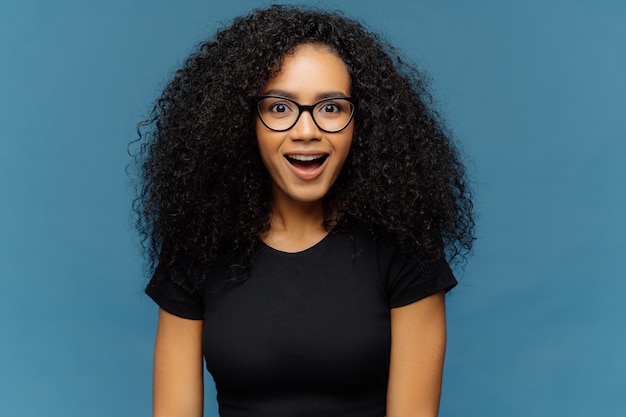 Angenehm überraschte, dunkelhäutige Frau, die mit Interesse die Kinnlade senkt, hat lockiges Haar, trägt schwarze T-Shirt-Modelle an der blauen Wand