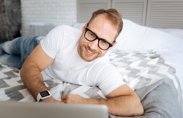 Angenehme Arbeitszeiten zu Hause. Erfreut lächelnder fröhlicher Mann, der auf dem Bett liegt und den Laptop benutzt, während er Interesse ausdrückt und arbeitet