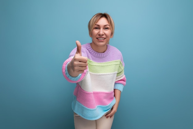 Foto angenehme blonde junge frau in einem gestreiften pullover zeigt eine super geste auf blauem hintergrund