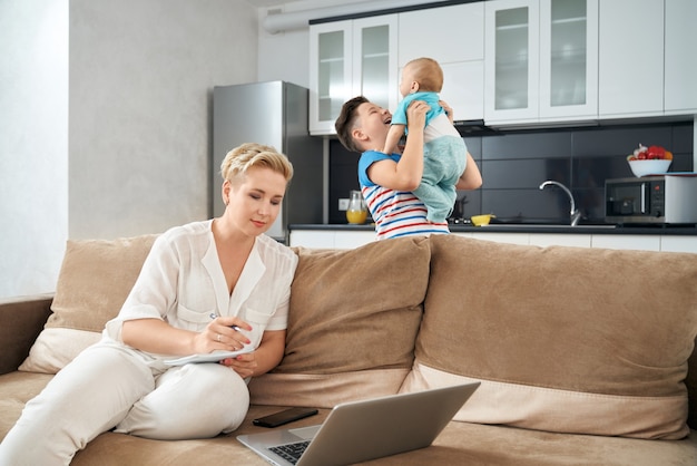 Angenehme Frau, die am Laptop arbeitet, während Söhne zusammen spielen