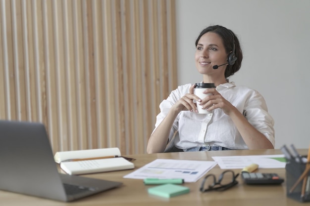 Angenehme italienische Geschäftsfrau im Headset sitzt am Schreibtisch im Heimbüro mit Kaffee zum Mitnehmen