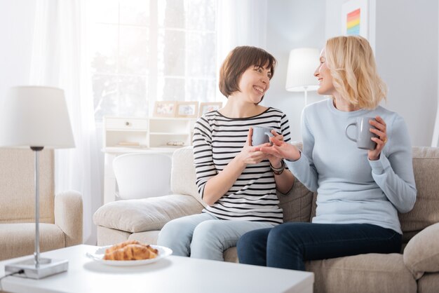 Angenehme Kommunikation. Nette glückliche entzückte Frauen, die zusammen sitzen und Tassen Tee halten, während sie ein angenehmes Gespräch führen