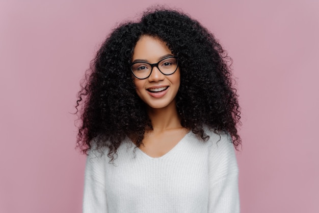 Angenehme schauende lächelnde Frau mit Afrofrisur, trägt optische Gläser und weiße Strickjacke