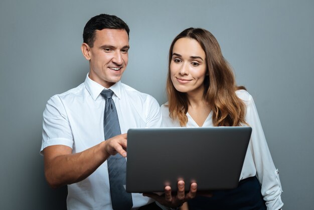 Angenehme Zusammenarbeit. Netter angenehmer fröhlicher Mann, der mit seiner Kollegin steht und ihr ein Bild zeigt, während sie einen Laptop hält