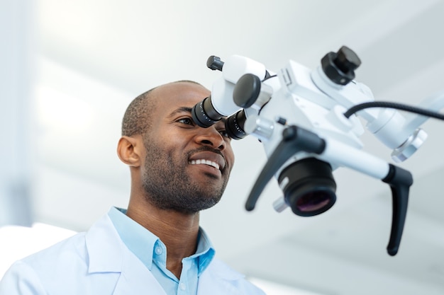 Angenehmer fröhlicher männlicher Zahnarzt, der breit lächelt und in die Linse eines Mikroskops schaut, während er die Zähne seines Patienten untersucht