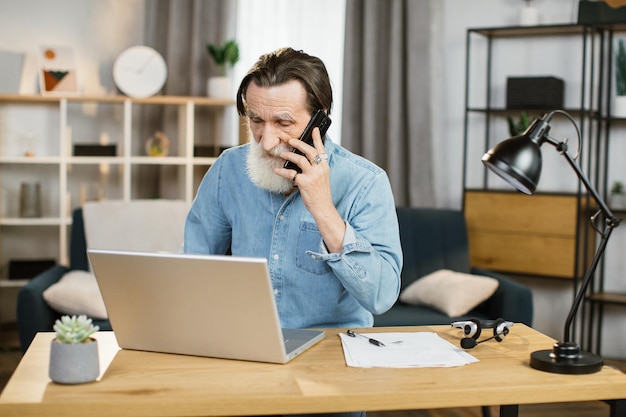 Angenehmer, reifer, kaukasischer Geschäftsmann, der am Handy spricht und am Tisch am Laptop arbeitet