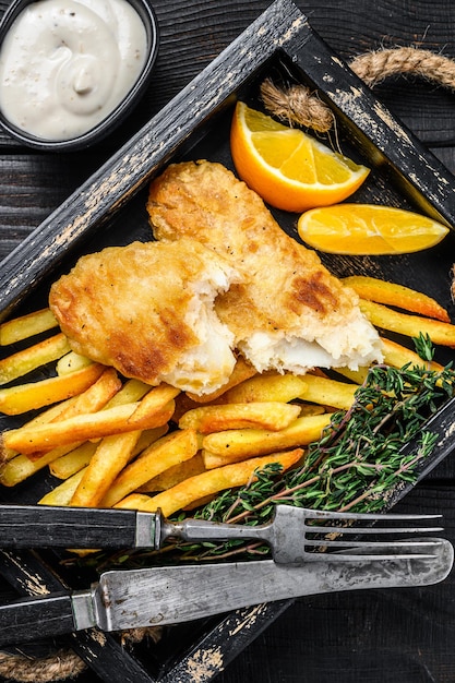 Angeschlagener Fisch und Pommes-Gericht mit Pommes frites und Remoulade in einem Holztablett. Schwarzer hölzerner Hintergrund. Ansicht von oben.