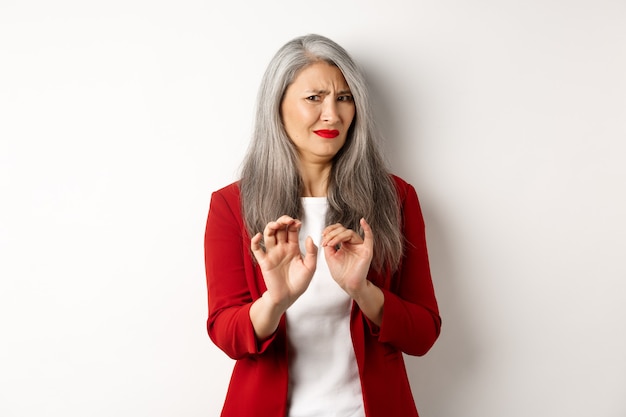 Angewiderte asiatische Geschäftsfrau mit grauem Haar, rotem Blazer und Make-up tragend, etwas Ekelhaftes ablehnend, Stoppschild zeigend, weißer Hintergrund.