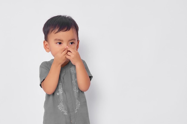 Angewiderter Kleinkind asiatischer muslimischer Junge, der sich angewidert fühlt und ihre Nase hält, um zu vermeiden, dass er auf weißem Studiohintergrund isoliert riecht