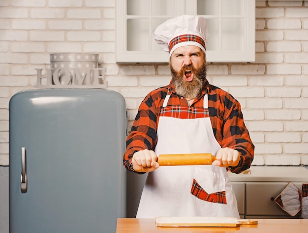 Angry chef panaderos hombre levantando rodillo amenazante en cocina blanca