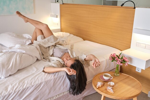 Foto Ángulo alto de mujer morena adulta en ropa de dormir elegante y bata de baño acostada en la cama boca abajo después de despertarse por la mañana con el desayuno en la mesita de noche