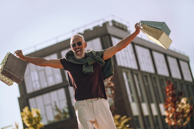 Ângulo baixo de um homem feliz com sacolas de compras nas mãos, posando para a câmera contra um prédio de escritórios