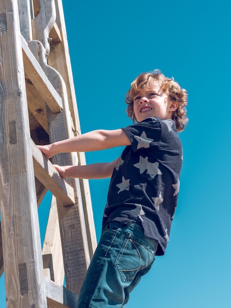 Ángulo bajo de niño rubio mirando hacia otro lado con una sonrisa mientras está de pie en una escalera de madera contra el cielo azul sin nubes