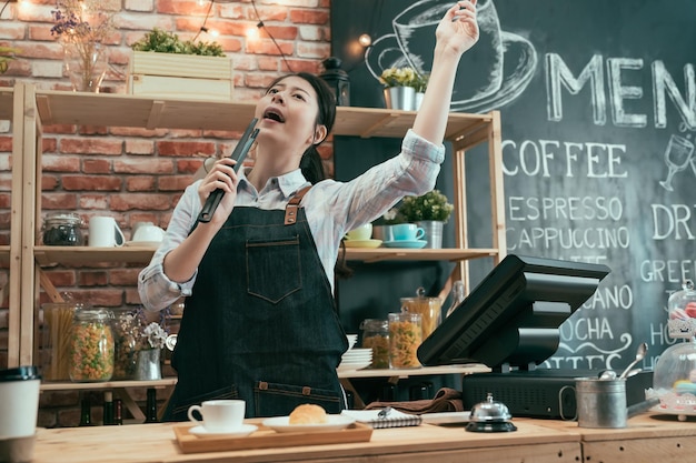 el ángulo bajo de la propietaria de una mujer de negocios en una cafetería disfruta de la música en el mostrador. camarera coreana asiática despreocupada de pie y bailando mientras canta con pinzas de pan en la cafetería después de preparar el pedido.