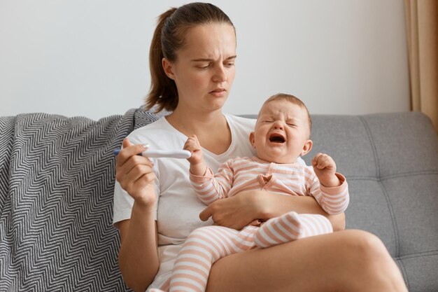 Angustiada mujer de cabello oscuro con coleta esperando el resultado de la prueba de embarazo pensando en el futuro mientras está sentado en el sofá en la sala de estar con la hija del niño en las manos.