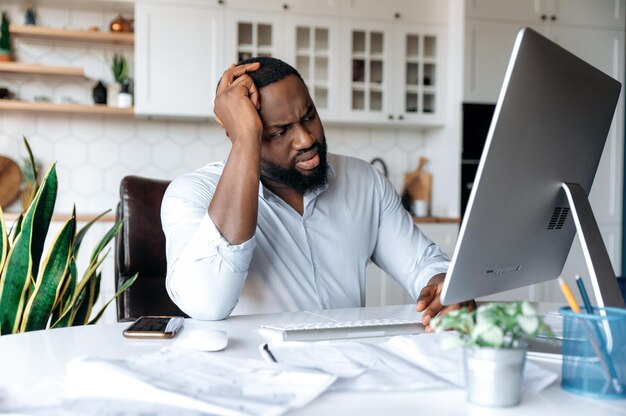 Angustiado triste joven afroamericano hombre gerente corredor o freelance trabajando en casa en la computadora experimentando estrés en el trabajo sosteniendo la cabeza con la mano mirando tristemente a la pantalla