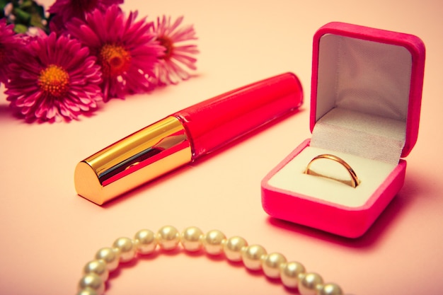 Anillo de oro en caja, abalorios, pintalabios y flores sobre fondo rosa. Joyas y complementos para mujer. Tonos de color.