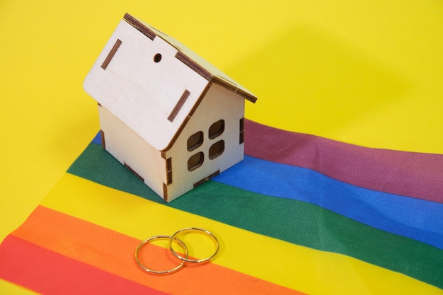 Anillos de boda y una pequeña casa de madera en la bandera lgbt, fondo amarillo, lugar de copia, vida familiar de familias del mismo sexo en un concepto de casa privada