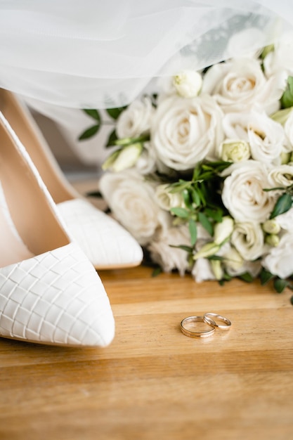 Foto anillos de boda, zapatos blancos y un ramo sobre una superficie de madera.