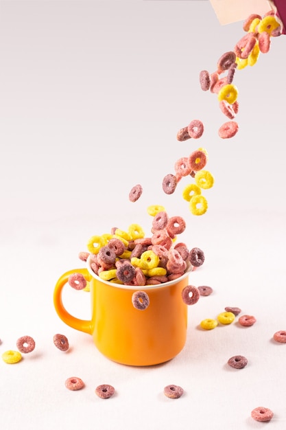 Anillos de cereales de colores cayendo en una taza naranja sobre fondo blanco.