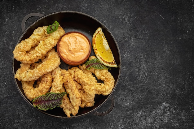 Anillos de oro de calamares fritos en una sartén redonda con salsa de hierbas y limón sobre un fondo oscuro