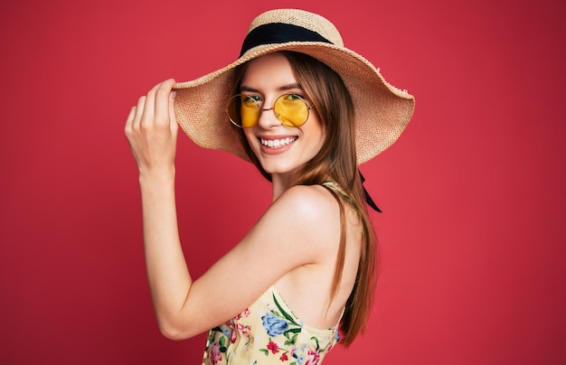 Animada e feliz linda jovem adorável em óculos de sol, vestido e chapéu de verão é se divertir enquanto posava em fundo rosa