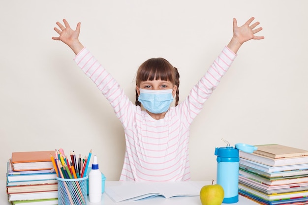 Animada feliz garotinha positiva com tranças usando máscara protetora sentada na mesa e levantou os braços cercada de livros e outros materiais escolares sendo feliz de volta à escola