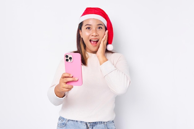 Animada jovem asiática com um chapéu de Natal segurando o telefone celular e segurando a bochecha isolada na celebração de fundo branco feriado de Natal e conceito de ano novo