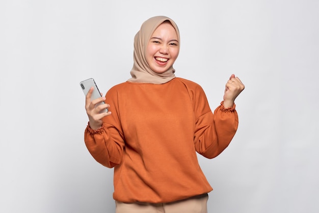 Animada jovem asiática de camisa laranja usando telefone celular e fazendo gesto de vencedor isolado sobre fundo branco