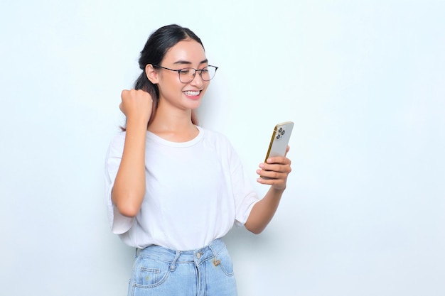 Animada jovem asiática em camiseta branca usando smartphone e fazendo gesto de vencedor isolado em fundo branco