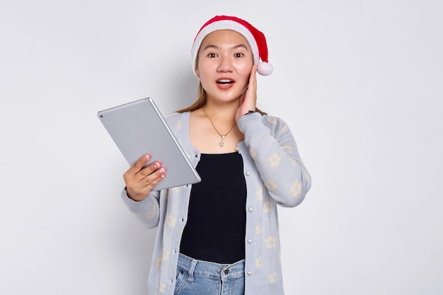 Animada jovem asiática em um chapéu de Natal usando um tablet digital e tocando sua bochecha isolada sobre fundo branco Povo indonésio celebra o conceito de Natal