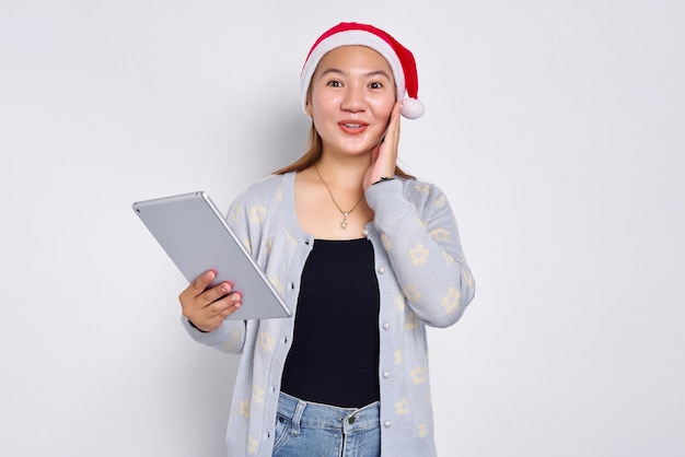 Animada jovem asiática em um chapéu de Natal usando um tablet digital e tocando sua bochecha isolada sobre fundo branco Povo indonésio celebra o conceito de Natal