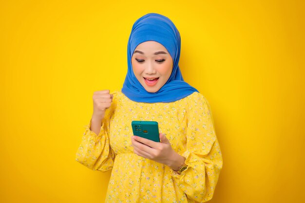 Animada jovem asiática em vestido casual usando telefone celular e fazendo gesto de vitória isolado em fundo amarelo