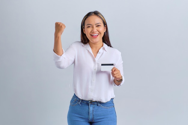 Animada jovem asiática segurando cartão de crédito e fazendo gesto de sucesso isolado sobre fundo branco