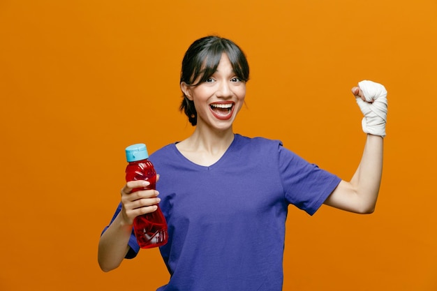 Animada jovem desportiva vestindo camiseta segurando a garrafa de água olhando para a câmera mostrando um forte gesto com o pulso envolto com curativo isolado em fundo laranja