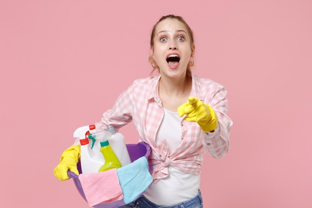 Animada jovem dona de casa em luvas de borracha segura bacia com garrafas de detergente lavando produtos de limpeza fazendo trabalhos domésticos isolados no fundo rosa. conceito de limpeza. apontando o dedo indicador na câmera.