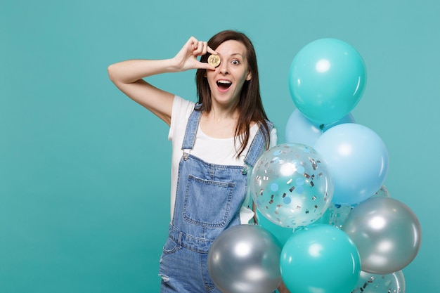 Animada jovem engraçada cobrindo os olhos com bitcoin, moeda futura, comemorando, segurando balões de ar coloridos isolados em fundo azul turquesa. Festa de aniversário, conceito de emoções de pessoas.