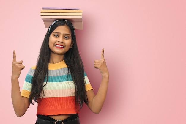 Animada jovem estudante asiática indiana posando livros de equilíbrio isolados na cabeça e apontando para cima com as duas mãos para um espaço de cópia olhando diretamente para a câmera expressando emoções positivas