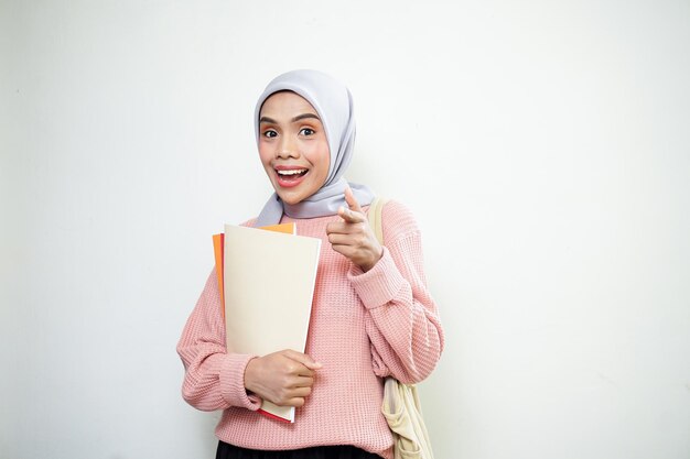 Animada jovem estudante muçulmana asiática em suéter rosa com bolsa e segurando livro