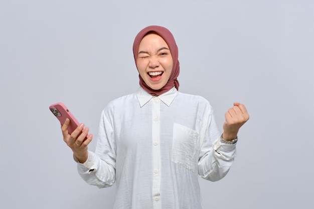 Animada jovem muçulmana asiática usando telefone celular e comemorando o sucesso recebendo boas notícias isoladas sobre fundo branco