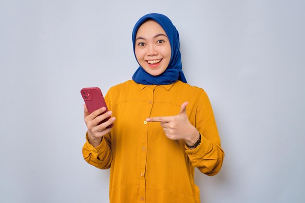 Animada jovem muçulmana asiática vestida de camisa laranja apontando dedos para o celular reagindo ao novo aplicativo isolado sobre fundo branco