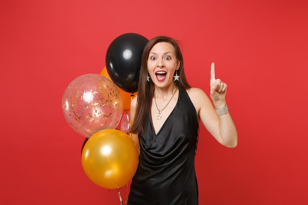 Animada jovem num vestido preto comemorando apontar o dedo indicador para cima segurando balões de ar isolados sobre fundo vermelho. Feliz dia internacional da mulher feliz ano novo, conceito de festa de feriado de maquete de aniversário.