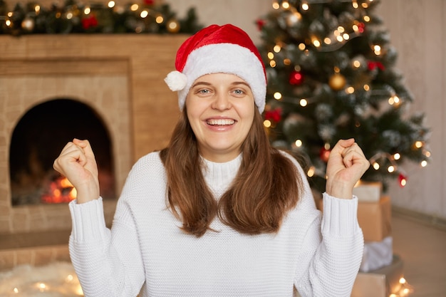 Animada jovem santa na camisola branca casual e chapéu de Natal cerrando os punhos, fazendo gesto de vencedor, celebração de ano novo, com sorriso encantador, estar feliz, tem bom humor.