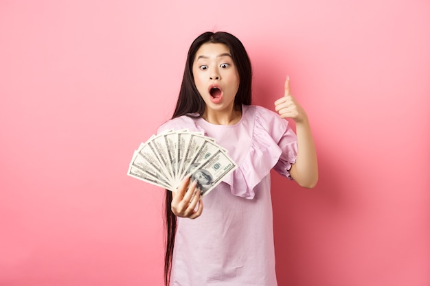 Animada menina adolescente segurando grande quantidade de dinheiro, mostrando notas de dólar e polegares para cima, em pé espantada no fundo rosa.