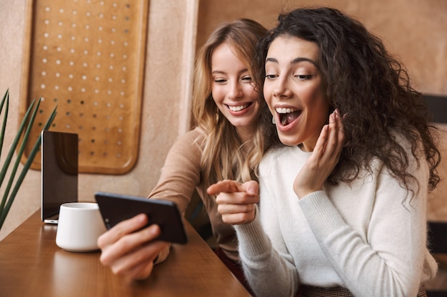 animadas amigas bonitas felizes sentadas em um café usando telefone celular