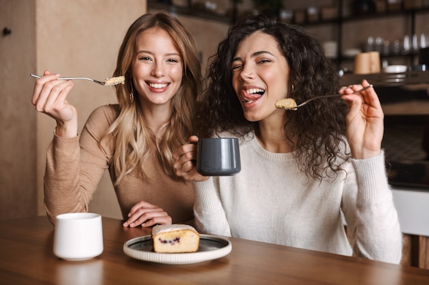 Foto animadas e felizes amigas lindas sentadas em um café tomando café e comendo bolo