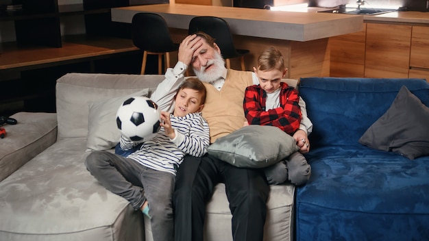 Animadas três gerações de fãs de esporte de homens relaxam na sala de estar comemoram a vitória do time juntos, menino muito feliz com o pai e o avô se divertem assistindo jogo de futebol em casa juntos